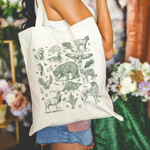 May include: A cream-colored canvas tote bag featuring a dark green screen-printed illustration of various North American wildlife, including a bear, deer, wolf, raccoon, owl, and other forest animals arranged in a collage pattern.