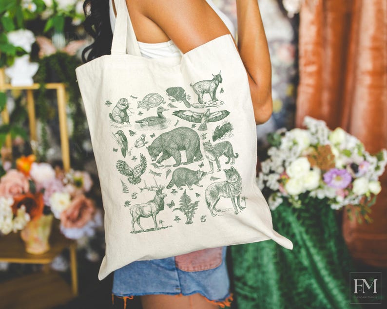 May include: A cream-colored canvas tote bag featuring a dark green screen-printed illustration of various North American wildlife, including a bear, deer, wolf, raccoon, owl, and other forest animals arranged in a collage pattern.
