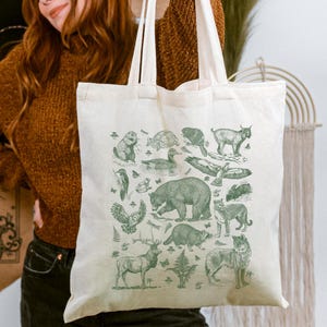 May include: A cream-colored canvas tote bag featuring a detailed green ink illustration of various North American wildlife, including a bear, wolf, raccoon, deer, owl, and eagle, arranged in a collage pattern with two long shoulder straps.