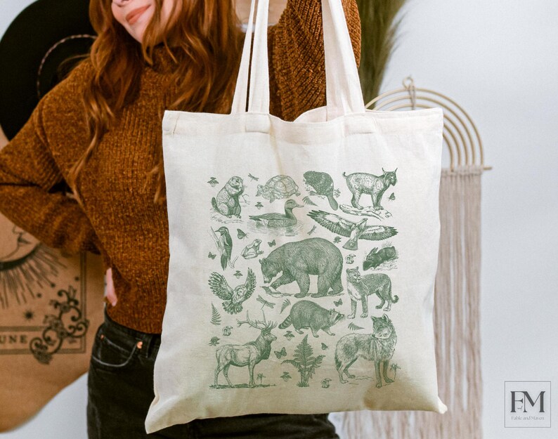 May include: A cream-colored canvas tote bag featuring a detailed green ink illustration of various North American wildlife, including a bear, wolf, raccoon, deer, owl, and eagle, arranged in a collage pattern with two long shoulder straps.