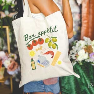 May include: A white canvas tote bag with the text "Bon app&eacute;tit" in green lettering. The bag features a colorful illustration of tomatoes, olives, pasta, a shrimp, and a can of sardines.