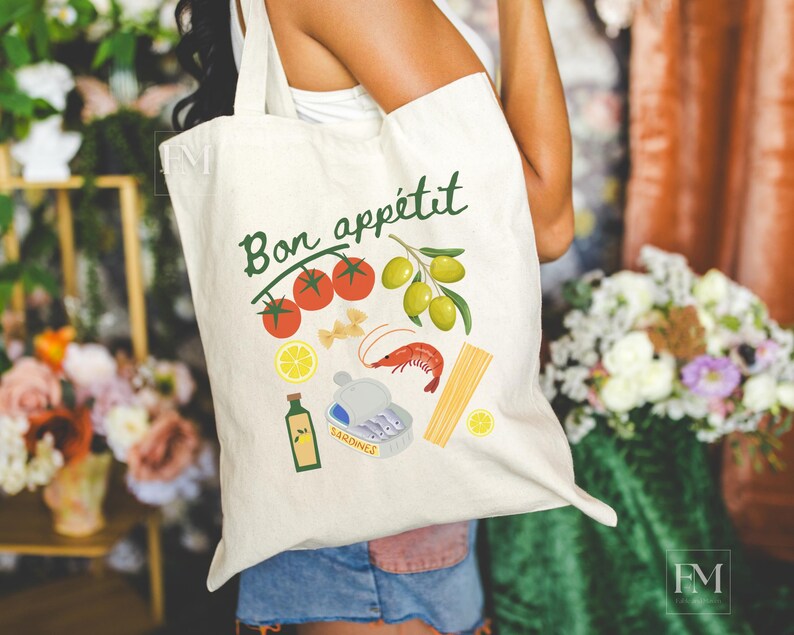 May include: A white canvas tote bag with the text "Bon app&eacute;tit" in green lettering. The bag features a colorful illustration of tomatoes, olives, pasta, a shrimp, and a can of sardines.