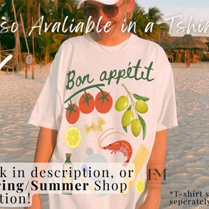 May include: White t-shirt with the text "Bon app&eacute;tit" in black lettering. The shirt features a graphic design of tomatoes, olives, pasta, a shrimp, and a lemon. The text "*T-shirt sold separately" is visible in the bottom right corner.