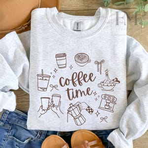 May include: Light gray sweatshirt with a brown graphic design featuring coffee-related illustrations and the words "coffee time." The design includes coffee cups, a coffee maker, and other coffee-themed elements. The sweatshirt is folded with a pair of blue jeans.