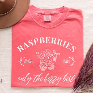 May include: A coral-colored t-shirt with the word "RASPBERRIES" in a white, arched font. Below the word are illustrations of raspberries and the phrases "HAND PICKED" and "FRESH DAILY". The text "only the berry best" is written in cursive.