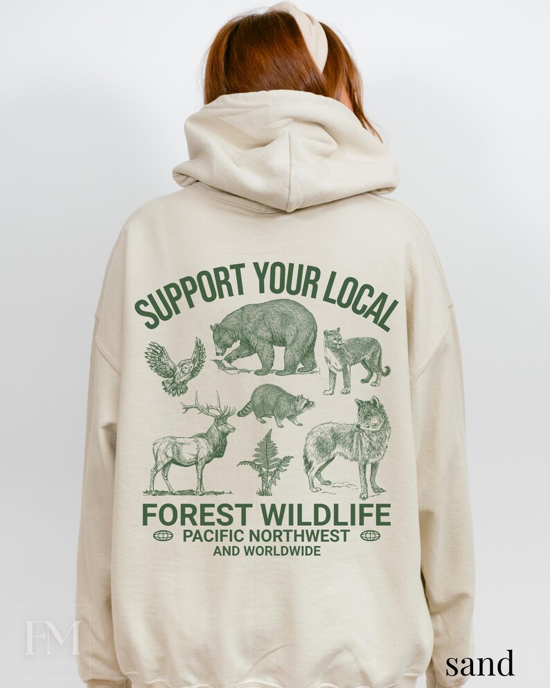 Puede incluir: Una sudadera con capucha beige con un estampado gr&aacute;fico verde que dice "Support Your Local Forest Wildlife Pacific Northwest and Worldwide". El gr&aacute;fico presenta un oso, un lobo, un ciervo, un b&uacute;ho, un mapache y un puma.