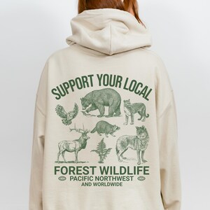 May include: A beige hoodie with a green graphic print that says "Support Your Local Forest Wildlife Pacific Northwest and Worldwide". The graphic features a bear, a wolf, a deer, an owl, a raccoon, and a cougar.