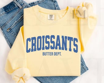 May include: A light yellow sweatshirt with the word "CROISSANTS" in blue block letters, with "BUTTER DEPT." underneath. The sweatshirt is laid on top of a pair of blue jeans.