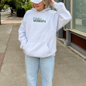May include: A gray hooded sweatshirt with the text "Collect the Moments" in green lettering. The model is wearing a white baseball cap and light blue jeans.