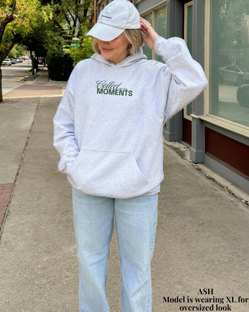 May include: A gray hooded sweatshirt with the text "Collect the Moments" in green lettering. The model is wearing a white baseball cap and light blue jeans.