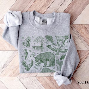 Pode incluir: Uma camisa de moletom cinza com uma estampa verde de v&aacute;rios animais, incluindo um urso, um veado, um p&aacute;ssaro e uma tartaruga. A camisa de moletom tem gola redonda e mangas compridas.