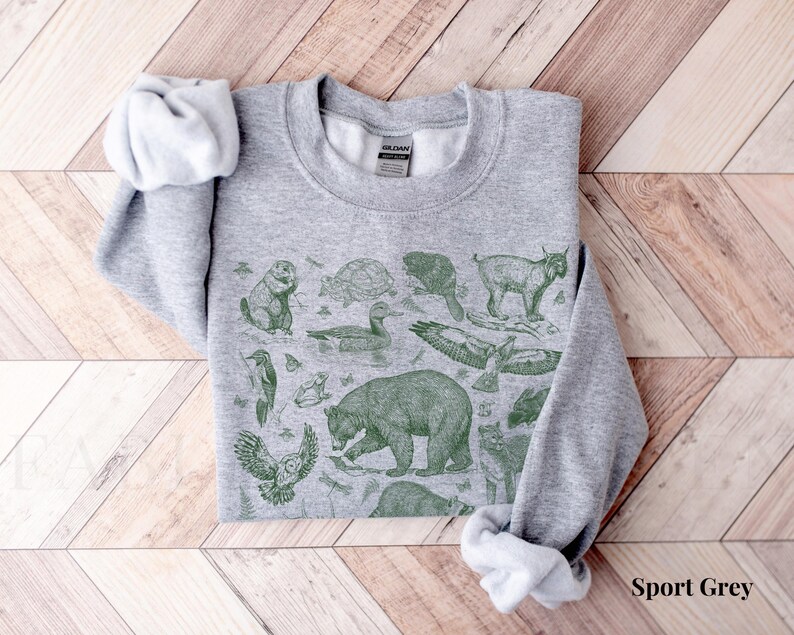 Pode incluir: Uma camisa de moletom cinza com uma estampa verde de v&aacute;rios animais, incluindo um urso, um veado, um p&aacute;ssaro e uma tartaruga. A camisa de moletom tem gola redonda e mangas compridas.