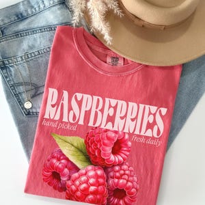 May include: A pink t-shirt with the text "RASPBERRIES hand picked fresh daily" in white. The shirt features a graphic of red raspberries with green leaves.
