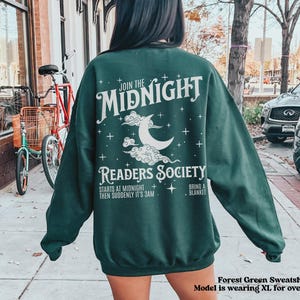 Może przedstawiać: Zielony sweatshirt z białym nadrukiem, na kt&oacute;rym widnieje napis "Join the Midnight Readers Society" z p&oacute;łksiężycem i gwiazdami. Tekst pod grafiką brzmi "Starts at midnight, then suddenly it's 3am. Bring a blanket."