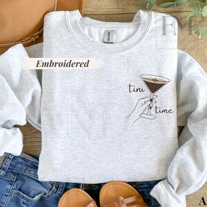May include: Light gray sweatshirt with the words "tini time" and a martini glass graphic. The sweatshirt is embroidered and has long sleeves. The image also shows a pair of blue jeans, sandals, and a brown hat.