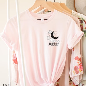 May include: A soft pink t-shirt with a black graphic of a crescent moon and clouds. The text "MIDNIGHT READING SOCIETY" is below the moon.