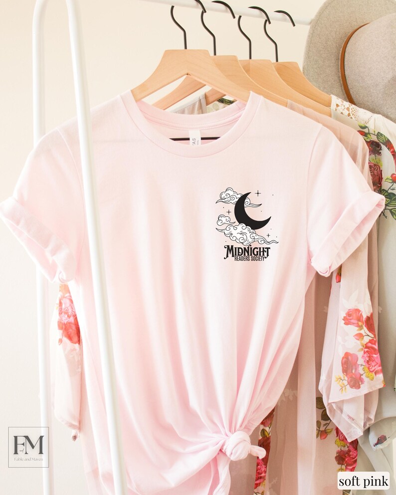 May include: A soft pink t-shirt with a black graphic of a crescent moon and clouds. The text "MIDNIGHT READING SOCIETY" is below the moon.