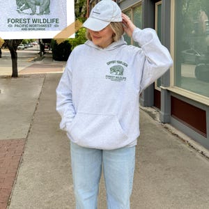 May include: A gray hooded sweatshirt with a green bear graphic and the text "Support Your Local Forest Wildlife Pacific Northwest and Worldwide".