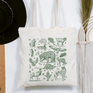 May include: A cream-colored canvas tote bag featuring a detailed green ink illustration of various North American wildlife, including a bear, deer, raccoon, wolf, owl, and eagle, arranged in a collage pattern with small ferns and butterflies.