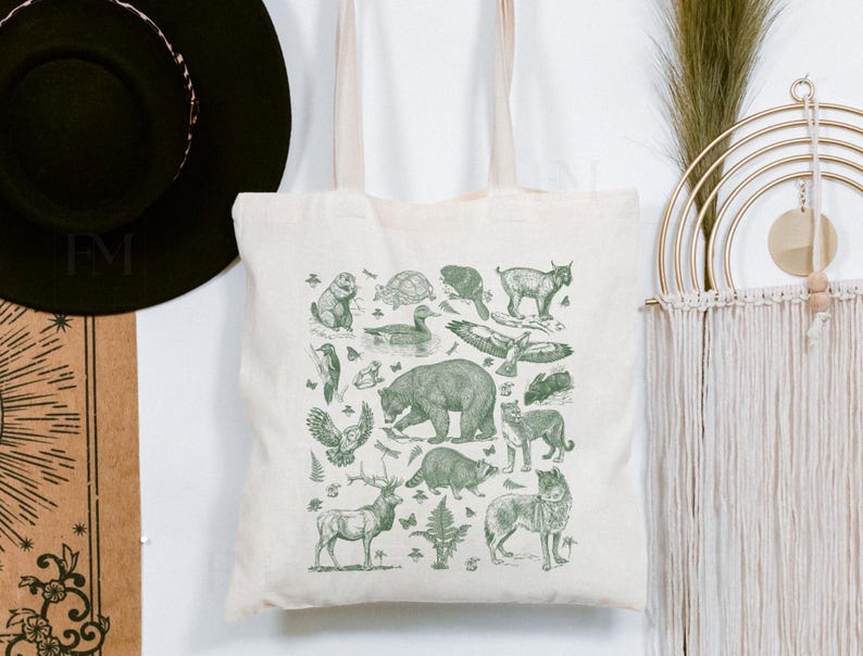 May include: A cream-colored canvas tote bag featuring a detailed green ink illustration of various North American wildlife, including a bear, deer, raccoon, wolf, owl, and eagle, arranged in a collage pattern with small ferns and butterflies.