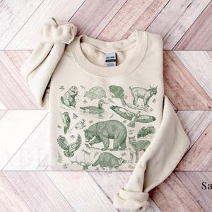 Pode incluir: Uma camisola bege com uma estampa verde de v&aacute;rios animais, incluindo um urso, um p&aacute;ssaro, uma tartaruga e um veado. A estampa &eacute; de estilo vintage e tem um toque r&uacute;stico. A camisola &eacute; feita de um tecido macio e confort&aacute;vel e &eacute; perfeita para o uso di&aacute;rio.