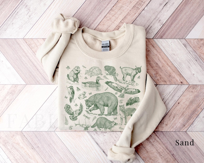 Pode incluir: Uma camisola bege com uma estampa verde de v&aacute;rios animais, incluindo um urso, um p&aacute;ssaro, uma tartaruga e um veado. A estampa &eacute; de estilo vintage e tem um toque r&uacute;stico. A camisola &eacute; feita de um tecido macio e confort&aacute;vel e &eacute; perfeita para o uso di&aacute;rio.