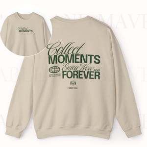 May include: A beige sweatshirt with long sleeves and a crew neck. The front of the sweatshirt has the text "Collect Moments Enjoy Now and Forever" printed in green. The back of the sweatshirt has the text "Collect Moments" printed in green.