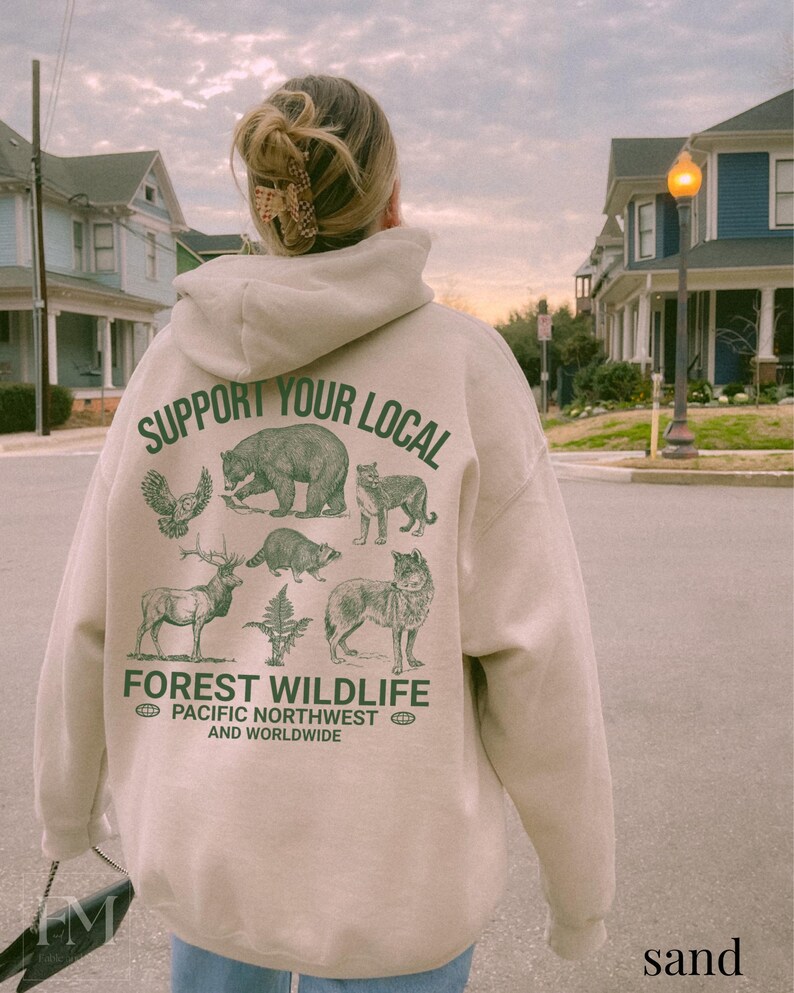 Puede incluir: Una sudadera con capucha beige con un estampado gr&aacute;fico verde de animales y el texto "Support Your Local Forest Wildlife Pacific Northwest and Worldwide".