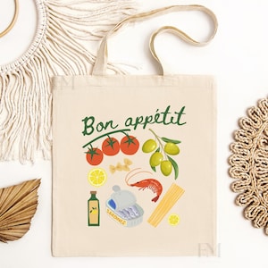 May include: A white canvas tote bag with the text "Bon app&eacute;tit" in green lettering. The bag features a colorful illustration of various food items, including tomatoes, olives, pasta, shrimp, lemons, and a bottle of sardines.