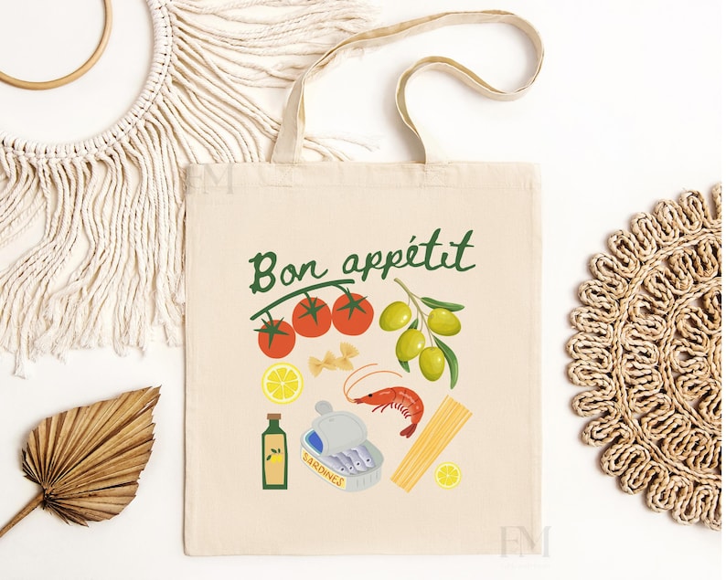 May include: A white canvas tote bag with the text "Bon app&eacute;tit" in green lettering. The bag features a colorful illustration of various food items, including tomatoes, olives, pasta, shrimp, lemons, and a bottle of sardines.