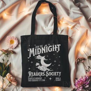 May include: Black tote bag with white text that reads "Join the Midnight Readers Society" with a crescent moon and clouds graphic. The text also says "Starts at midnight then suddenly it's 3 am. Bring a blanket."