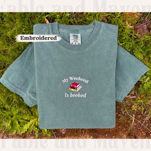 May include: A green t-shirt with the text "My Weekend Is Booked" embroidered on the front. The embroidery features a stack of books with a red spine.