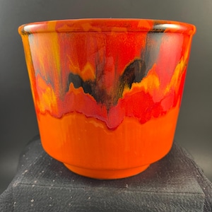 May include: A ceramic planter featuring a vibrant orange base that transitions into a glossy, multi-colored glaze at the top. The upper section displays swirling patterns of bright red, deep yellow, and black, creating a bold, fluid aesthetic.