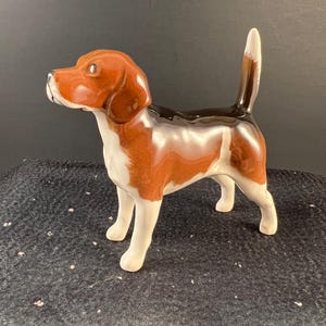 May include: A ceramic figurine of a beagle dog, standing with its tail up. The dog has a brown head and ears, a white chest and legs, and a black and brown back. The figurine is glossy and detailed.