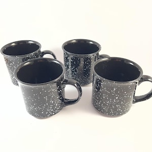 May include: A set of four black mugs with a white speckled pattern. Each mug features a cylindrical body, a smooth interior, and a curved handle on the side. The exterior surface of each mug is glossy and covered in small, irregular white dots.