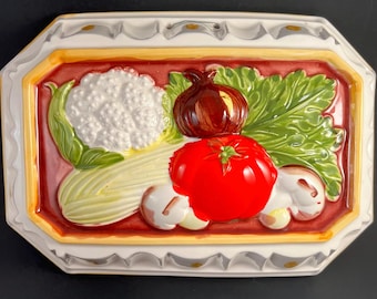 May include: A rectangular ceramic mold featuring a raised, colorful relief of vegetables including a white cauliflower, a red tomato, a brown onion, and green celery leaves against a deep red background with a yellow and white scalloped border.