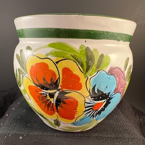 May include: A white ceramic planter featuring a hand-painted floral design with large yellow, orange, and blue pansy-like flowers and green leaves. The rim of the planter is accented with a solid green horizontal band around the top edge.