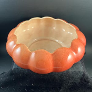 May include: A round ceramic bowl with a scalloped, pumpkin-like edge. The exterior features a glossy, vibrant orange glaze, while the interior is finished in a smooth, light cream color. The bowl has a wide, open top and a curved, segmented rim.