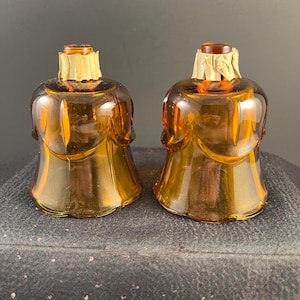 May include: Two amber-colored glass candle cups with a bell-shaped design and scalloped edges. Each cup features a narrow neck wrapped in a light tan paper collar at the top opening, showcasing a translucent finish and a smooth, curved glass body.