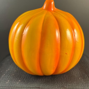 May include: A smooth, rounded glass pumpkin sculpture featuring a vibrant gradient of yellow and orange hues. The object has a glossy, reflective surface with vertical ridges that mimic the natural shape of a pumpkin, topped with a small stem.