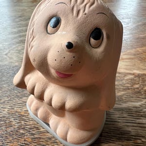 May include: A light tan, matte-finish ceramic figurine of a puppy with large, dark, round eyes, a small black nose, and a pink mouth. The puppy is depicted in a standing position with its front paws held together near its chest, featuring textured fur details on its head and ears.