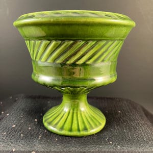 May include: A green ceramic pedestal bowl with a glossy finish. The bowl has a wide, fluted base and a decorative band of diagonal ridges around the middle. The rim has small holes. A vintage decorative piece.