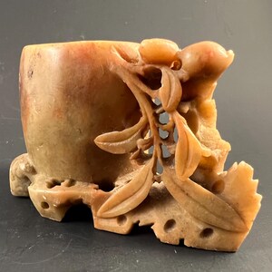 May include: A carved stone planter with a rectangular body and intricate floral and fruit details. The planter is a warm, earthy tone with variations of tan and orange. The design includes leaves, fruit, and openwork patterns, suggesting a decorative or artistic purpose.