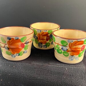 May include: Three cream-colored ceramic cups featuring a hand-painted floral design with large orange flowers, green leaves, and small blue accents. Each cup has a subtle crackle glaze finish and a brown rim, with a tapered cylindrical shape.