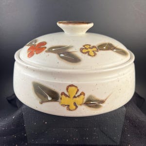 May include: A round ceramic casserole dish with a matching lid, featuring a speckled off-white glaze. The exterior and lid are decorated with hand-painted yellow and orange flowers accompanied by dark green leaves, finished with a brown knob.
