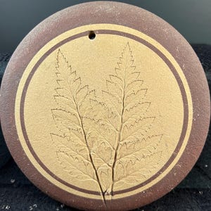 May include: A circular brown ceramic ornament featuring a central tan-colored disc with an embossed fern leaf design. The ornament has a dark brown border and a small hole at the top for hanging. The surface has a matte, textured finish throughout.
