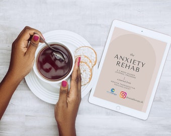 May include: A white tablet with a pink background and the text "the ANXIETY REHAB A 4 WEEK GUIDED JOURNAL PROGRAM a journal from KRISTIN THORISDOTTIR PSYCHOLOGIST @mindtherapy.uk" on the screen. The tablet is on a white wooden surface with a cup of tea and cookies.