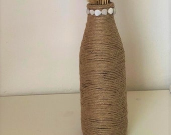 Op de afbeelding: Een bruine glazen fles omwikkeld met touw met een decoratieve band van witte kralen. De fles is gevuld met gedroogd pampasgras.