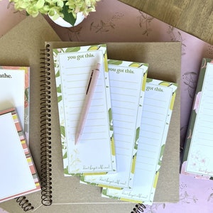 May include: Three white notepads with green and yellow floral designs and the text "you got this" and "got this". The notepads have a pink pen on top of them and the text "Don't forget self care!" at the bottom.