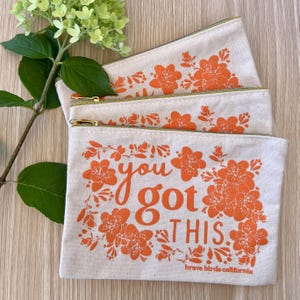 May include: Three canvas zipper pouches with orange floral designs and the phrase "you got this." The pouches have gold zippers and green trim. The text "brave birds california" is printed at the bottom of the bottom pouch.